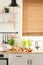 Real photo of bright kitchen interior with fresh plants, tomatoes and bread placed on countertop, window with wooden blinds and pa