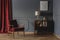 Real photo of a corner of a retro living room interior with elegant, beige lamp on a wooden cabinet next to a red armchair