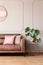 Real photo of end table with decor and fresh plant placed next to leather sofa with pink cushions in light grey living room interi