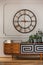 Real photo of a large, metal clock hanging on a white wall with molding above a wooden cupboard in bright living room interior