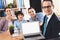 Realtor sitting at desk in office. Realtor is presenting laptop with family in background.