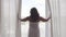 Rear back view at rich mature woman opening curtain lace standing in luxury apartment home or modern hotel looking