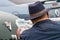 Rear shot of a caucasian man in a hat sat beside classic cars reading adverts for classic cars in a newspaper.  Over shoulder shot