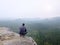 Rear view of alone hiker in dark outdoor suit sit on the edge. Sharp rocky peak above mountains valley