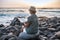 Rear view of alone senior woman with  hat sitting on the beach of pebbles reading a book. Waves of the ocean. Sunset light