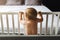 Rear view. A child alone is standing at home in a crib, holding on to its side and looking at an empty bed in the background