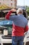 Rear view at confused Caucasian mature man scratching the back of his head with wrench while standing behind opened hood of car,