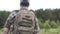 Rear view of fisherman in camouflage clothes with backpack walking with fishing poles in field to river. View from back