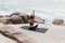 Rear view of fit female raising hand while changing yoga pose. Woman sitting on mat by shore and practicing yoga