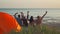 rear view four best friends sitting at sunset on beach in front of sea and raising hands