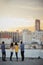 Rear View Of Friends On Rooftop Terrace Looking Out Over City Skyline At Sunset