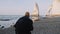 Rear view happy tourist man with backpack walking towards famous white arch cliff near Etretat in Normandy slow motion.