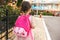 Rear view image of cute little girl preschooler walking outdoor with pink backpack against blurred buildings. Happy kid toodler