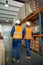 Rear view of industrial workers in white hardhat and protective vest inspecting the goods in the warehouse