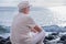 Rear view of senior caucasian man sitting on the pebble beach relaxing and looking at horizon over water. Elderly white haired man