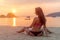 Rear view of slim female model sitting on seashore wearing bikini looking away during sunrise with sun path reflected