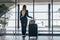 Rear view of slim young female traveler standing in airport terminal holding heavy suitcases looking through the window