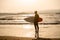 Rear view of strong surfer with surfboard on the beach at sunset or sunrise. Silhouette of surf man standing looking at ocean