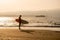 Rear view of strong surfer with surfboard on the beach at sunset or sunrise. Silhouette of surf man standing looking at ocean