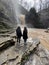 Rear view of two women looking on powerful muddy waterfall. Tourists standing on rocks in mountainous terrain and