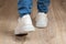 Rear view of walking feet shod in white chunky sole sneakers and blue jeans on the brown floor. Pair of new comfortable shoes for