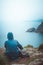 Rear view of woman sitting on the edge of a cliff with blue raincoat and backpack looking at the horizon and sea