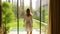 Rear view of young attractive woman standing near big full length window, touching glass. Fit female enjoying garden view.