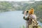 Rear view of young female backpacker in warm casualwear taking photo of river