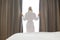 Rear view of young woman in bathrobe opening window curtains at hotel room