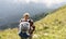 Rear view of young woman in cap and plaid shirt putting on backpack hiking in green mountains in summer healthy active lifestyle,