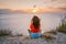 Rear view a young woman in a red shirt is sitting on the edge of a cliff in front of the sea with a delightful sunset