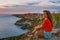 Rear view a young woman in a red shirt is sitting on the edge of a cliff in front of the sea with a delightful sunset