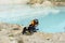 Rear view of young woman traveler in cap and ginger t-shirt stroking fluffy gray dog on sandy shore of blue lake or river, pet