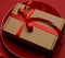 Rectangular brown cardboard box tied with a silk red ribbon lies in a round ceramic red plate