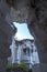 A rectangular opening in a dark, mossy limestone cave frames a bright blue sky with fluffy white clouds in Setigi