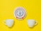 A red alarm clock on a saucer and two coffee cups on a yellow background. Flat lay.