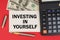 On a red background, among the money, a calculator and a pen lies a sign with the text - INVESTING IN YOURSELF