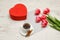 Red box in heart shape, pink tulips, gray sheet and a coffee mug. Light table