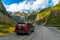 Red cars parked on a gravel road into the entrance to Fox Glacier, high valleys and blue skies with beautiful clouds