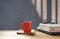 Red coffee cup and eyeglasses on stacked books, black laptop on wooden table in front of loft cement wall background