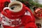 A red cup of hot coffee, a red and white knitted scarf with pompons, a fir twig, cones and Christmas-tree decorations