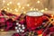 Red cup with hot coffee and sweet marshmallows on wooden table with plaid in Christmas decorations with garland lights