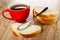 Red cup with tea, spoon on sandwich with condensed milk, slices of bread on wooden table