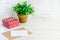 Red dotted gift box, empty card, kraft envelope and a green flower in a rustic ceramic pot. White wooden background, copy space.