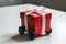 Red gift box with black wheels on white surface, ribbon pops against red.