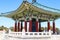 A red and green Korean Pavilion with massive bronze bell in the center with stone stairs on top of a hill with blue sky