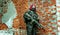 Red-hair airsoft woman in uniform with machine-gun, stand beside brick wall