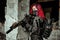 Red-hair airsoft woman in uniform with machine gun stand beside column. Closeup soldier stand inside of broken building