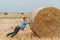 Red-haired boy pushing a bale of straw in the middle of a wheat field with bales on a summer evening
