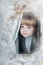 Red-haired charming girl looks out the window and smiles. Winter snow-covered window and blue-eyed girl looking at the camera